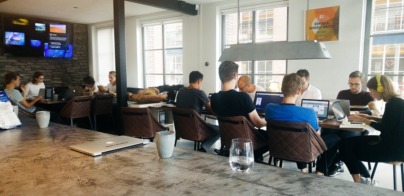 Open-plan coworking space with a long table of people working on laptops, brick wall with screens, large windows, and a hanging light.