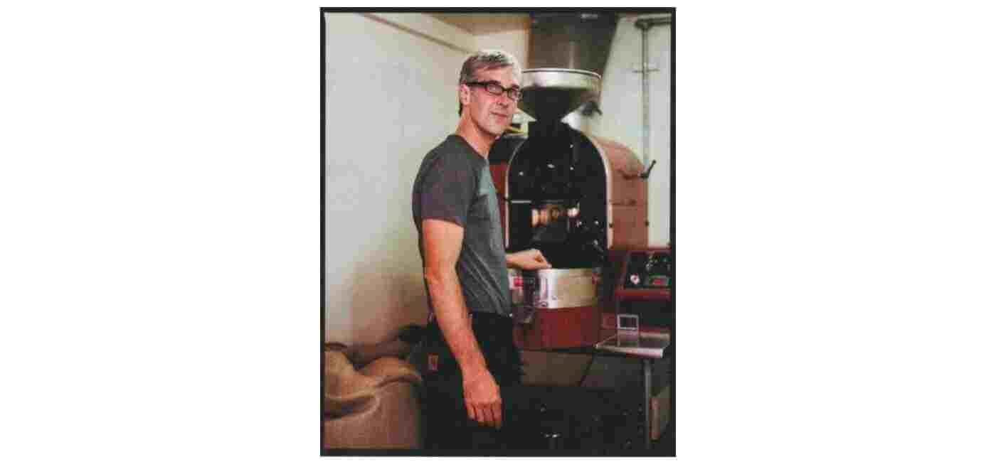 A person with short gray hair, wearing a gray t-shirt and glasses, stands beside a large industrial coffee roaster in a workshop.