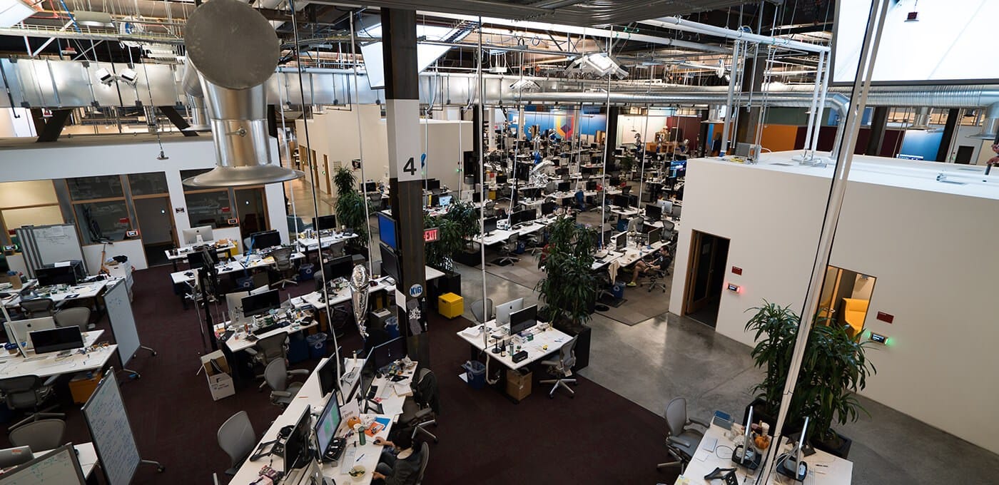 Open-plan office with numerous desks, computer monitors, and staff at work among exposed ductwork, pipes, and industrial lighting.