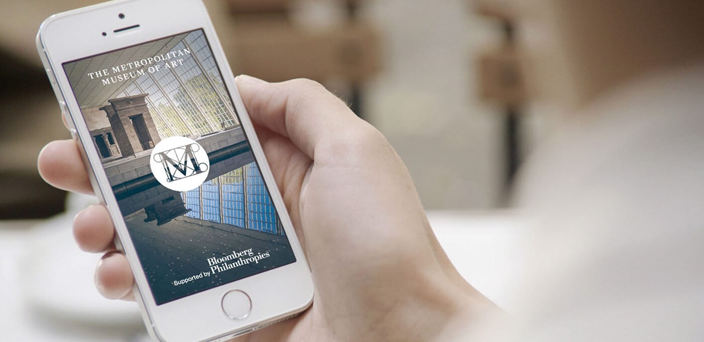 Hand holding a white iPhone displaying a poster for The Metropolitan Museum of Art with a Bloomberg Philanthropies logo.