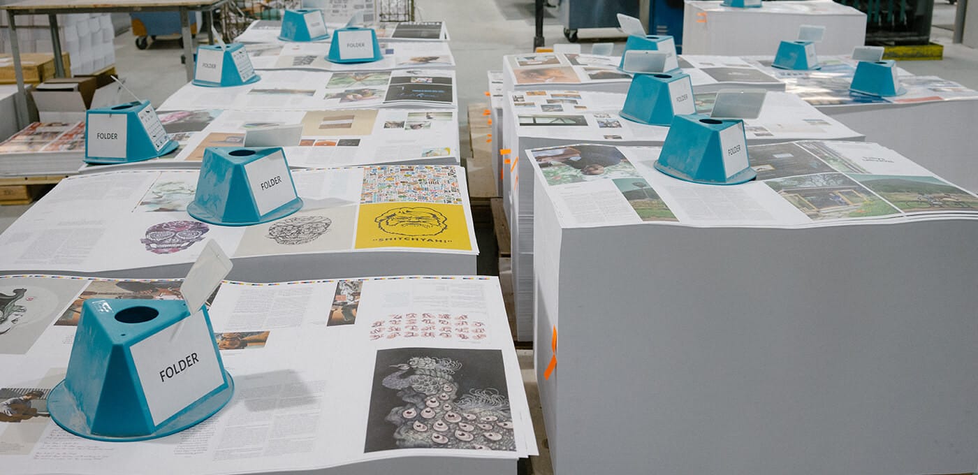Long rows of tables covered with magazines and pages, blue folder holders labeled FOLDER sit along the surfaces in a busy printing/archive space.