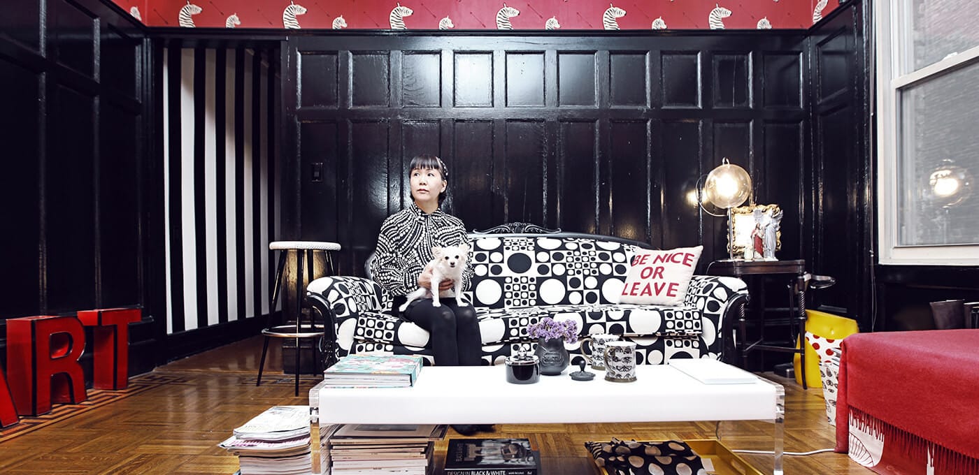 A person sits on a black-and-white geometric sofa in a living room, holding a small white dog; a coffee table with magazines sits in front.