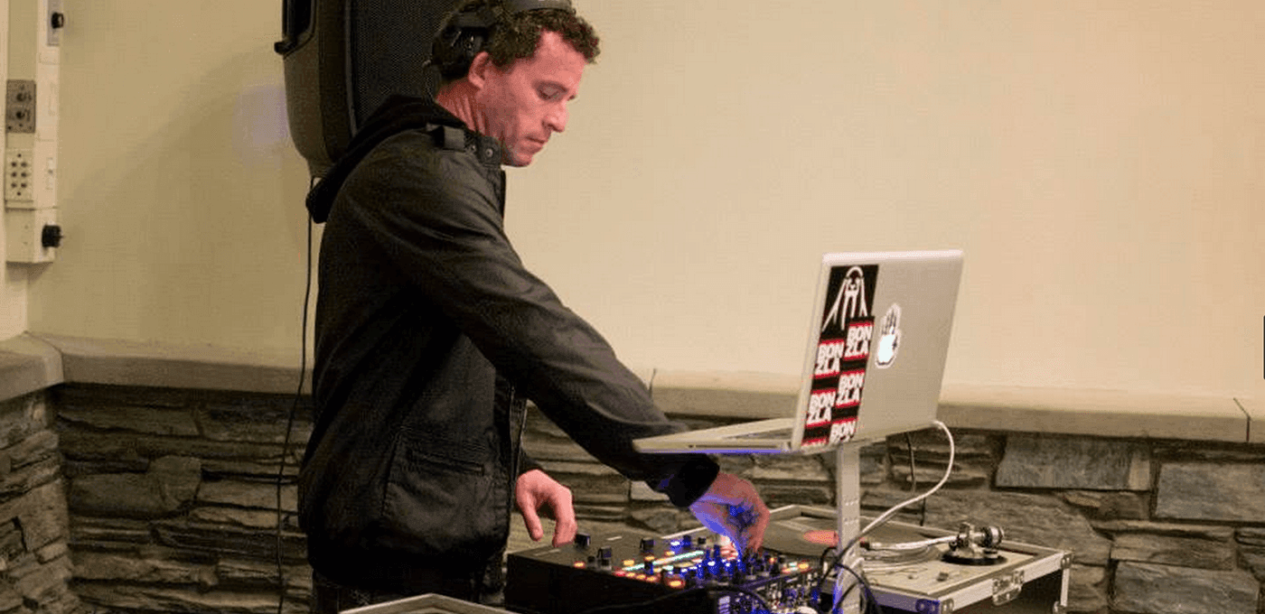 A DJ in a dark jacket wearing headphones adjusts a mixer, with a laptop on a stand nearby; a beige-walled room with a stone baseboard behind them.