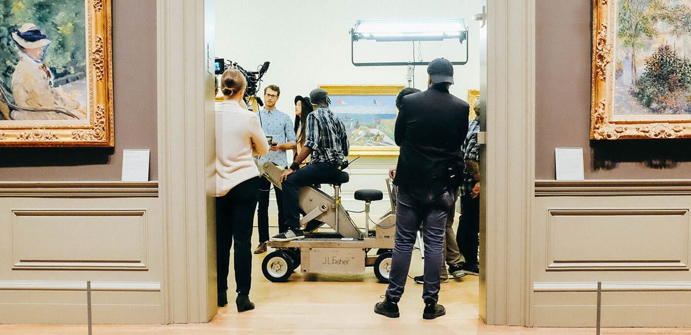 Film crew on a camera dolly in an art gallery, with bright overhead lights and framed paintings on the walls as onlookers watch.