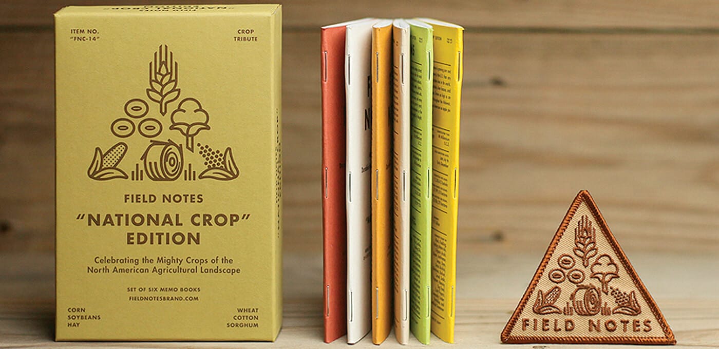 Yellow Field Notes National Crop Edition box beside a row of colorful notebooks and a stitched triangular Field Notes patch on a wooden surface.