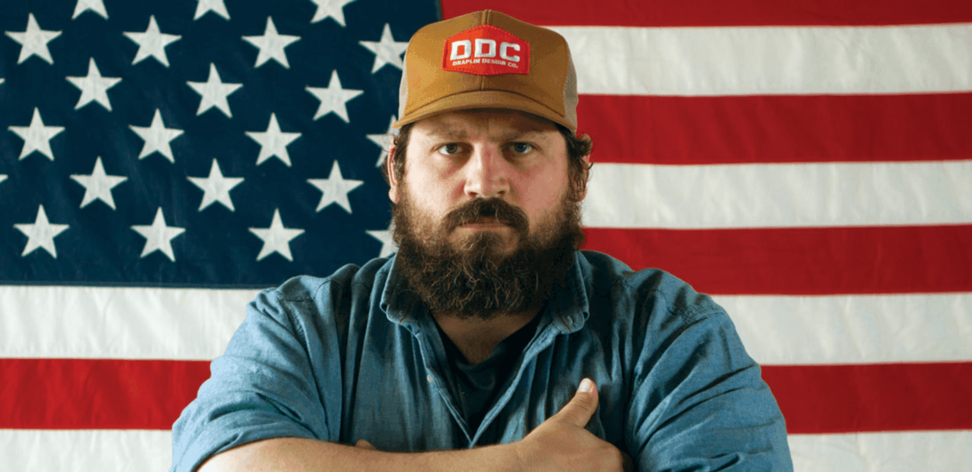 Bearded person in a blue denim shirt and brown cap with a patch stands in front of a large American flag, arms crossed, facing the camera.