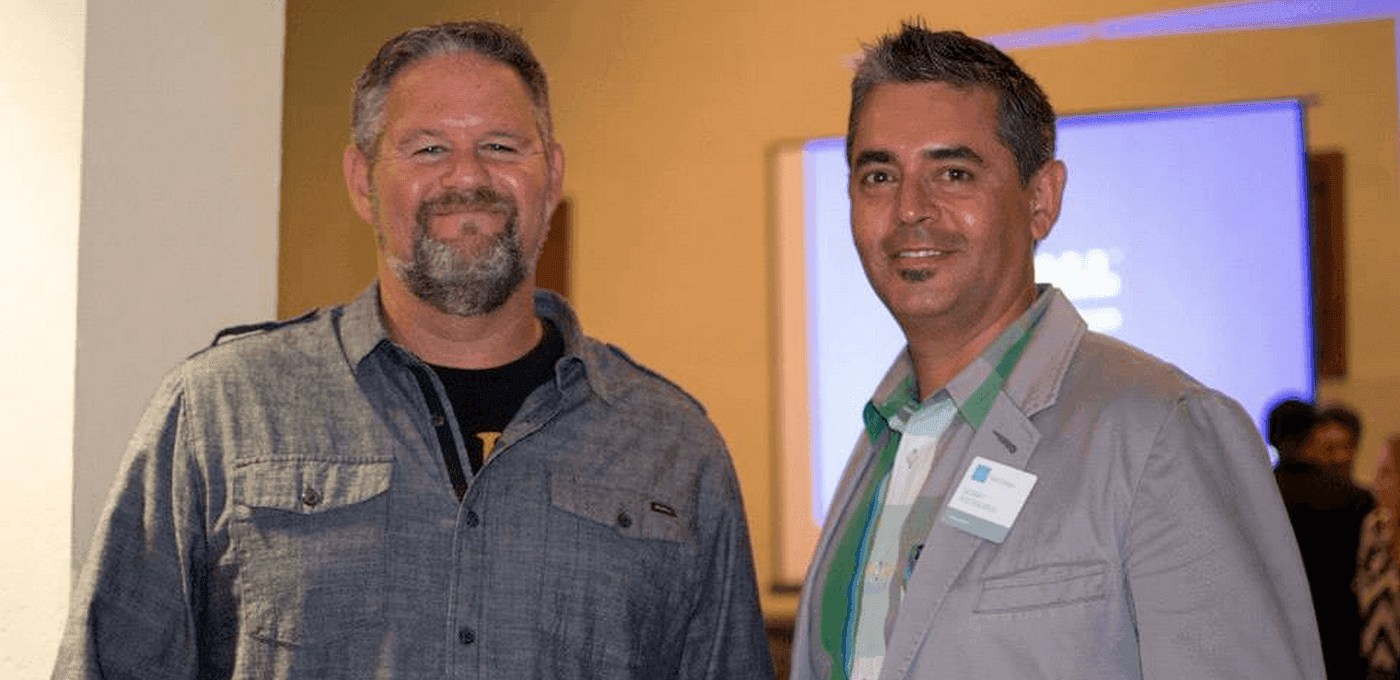 Two people indoors, smiling. Left person in a gray denim shirt with a beard; right person in a light gray blazer with a name badge, against a yellow wall.