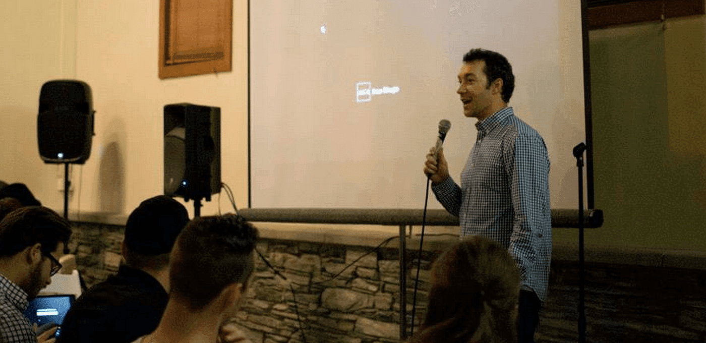 A person in a checkered shirt speaks into a microphone to an audience, standing by a projection screen with a Windows desktop icon.