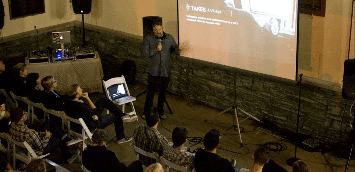 A speaker with a microphone shows a projected slide to an audience seated on white folding chairs; a laptop sits on a chair and audio gear nearby.