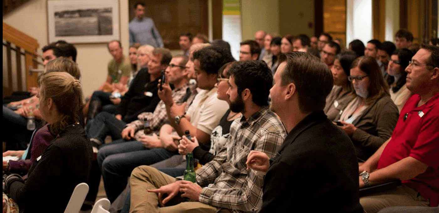 Diverse audience seated in rows inside a conference room, attentively watching a talk; some hold drinks or smartphones.