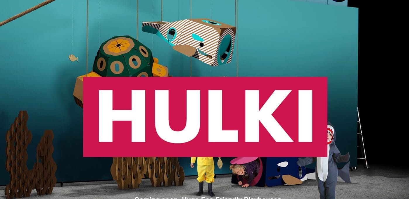 Teal stage backdrop with suspended cardboard sculptures and a bold pink "HULKI" sign across the foreground; costumed performers and large props at the bottom.