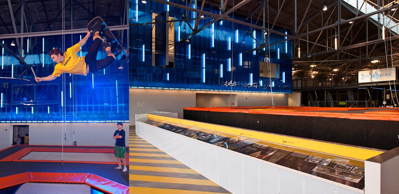 Indoor trampoline park: a person in a yellow shirt soars midair performing a trick on a trampoline, while another person watches; blue neon walls.