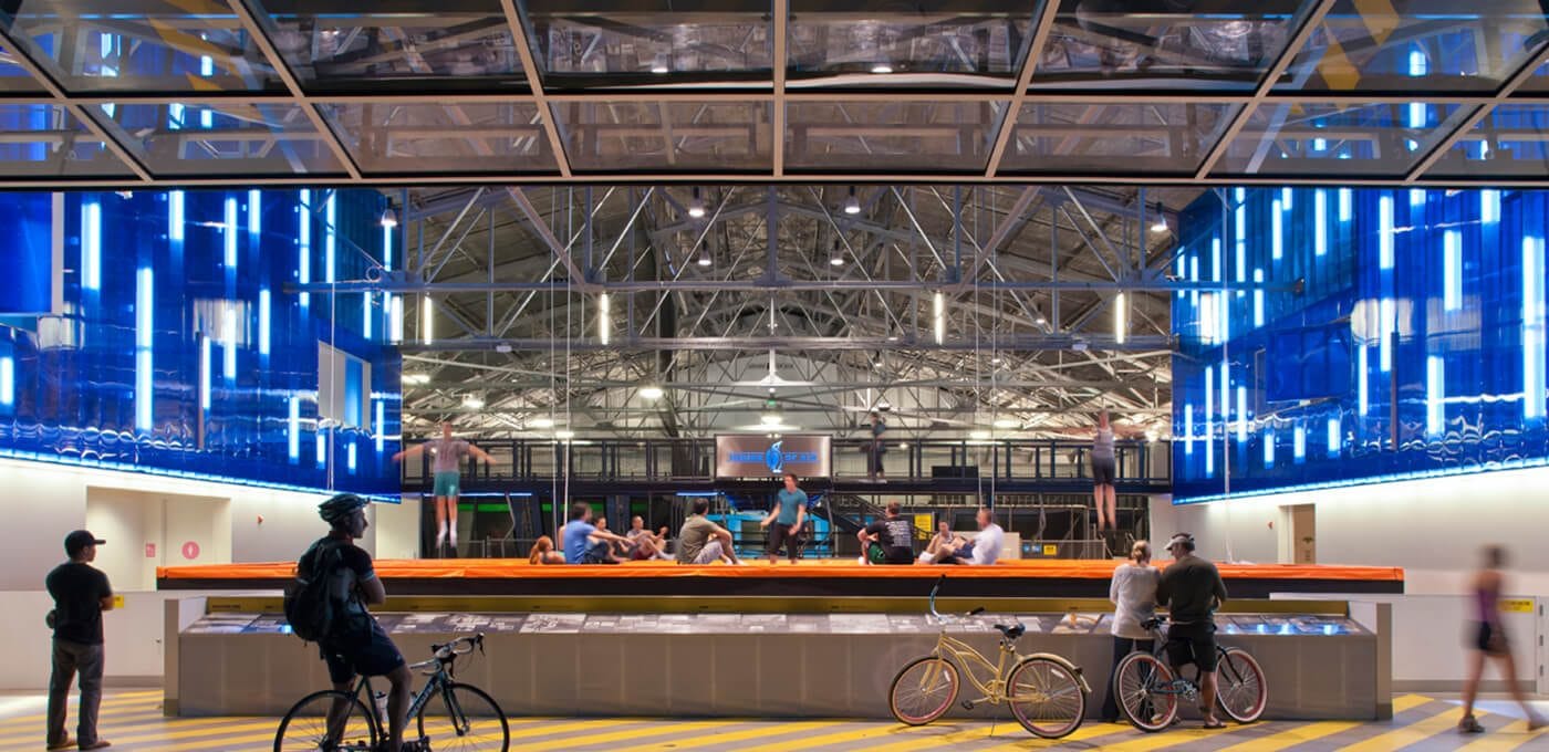 Indoor trampoline park with blue neon walls and a metal ceiling; people bounce on orange trampolines, while bikes and spectators stand nearby.