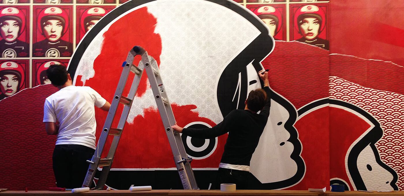 Two artists painting a red, black, and white mural with bold circular shapes; a ladder between them, background with repeating portrait posters.