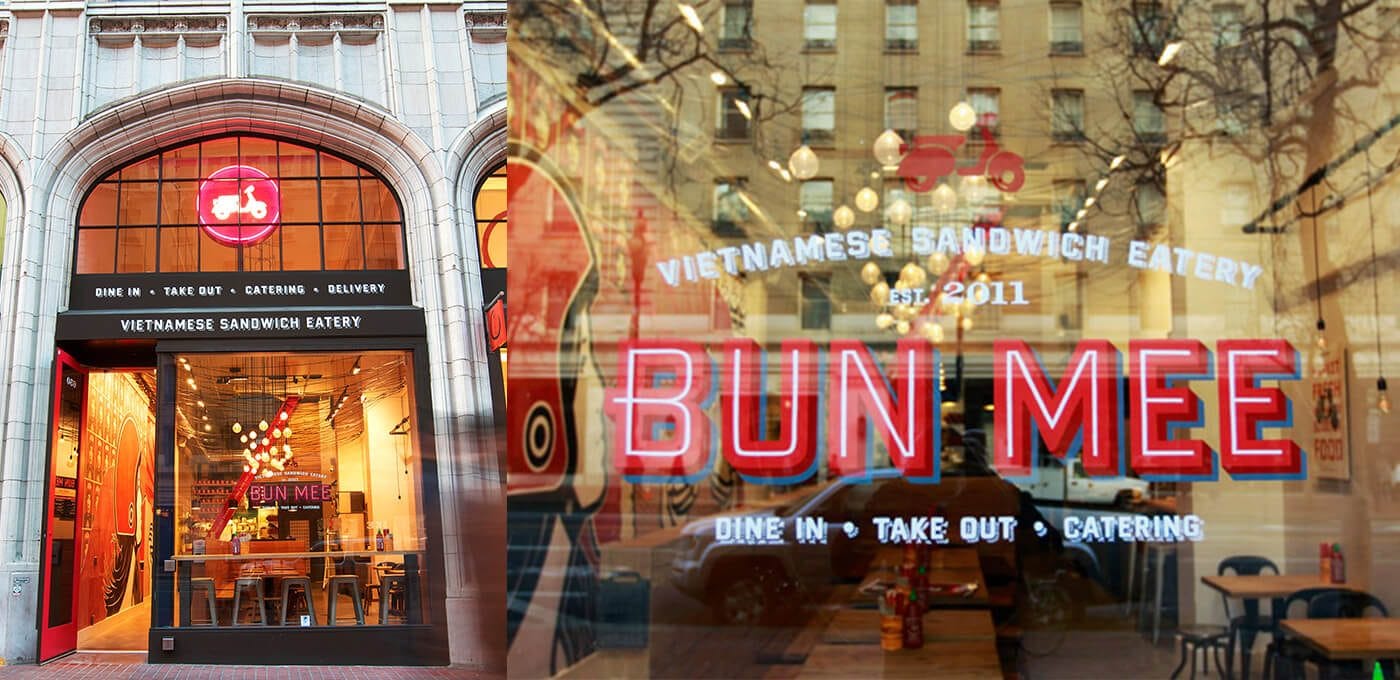 Front of a Vietnamese sandwich shop Bun Mee with an arched storefront, large windows, and a red neon BUN MEE sign; interior seating visible inside.