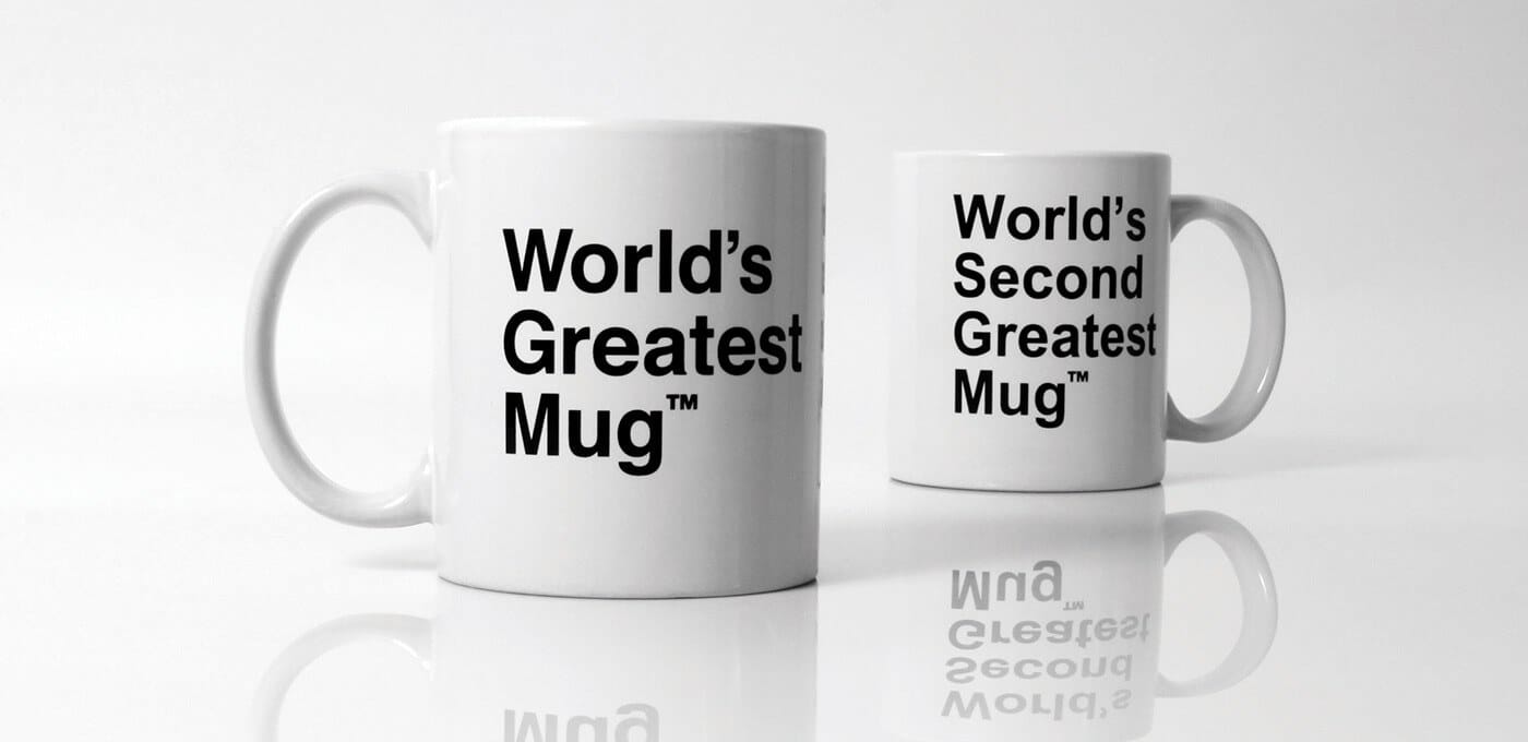 Two white mugs on a reflective surface; the left mug reads "World's Greatest Mug" and the smaller right mug reads "World's Second Greatest Mug" in bold black text.