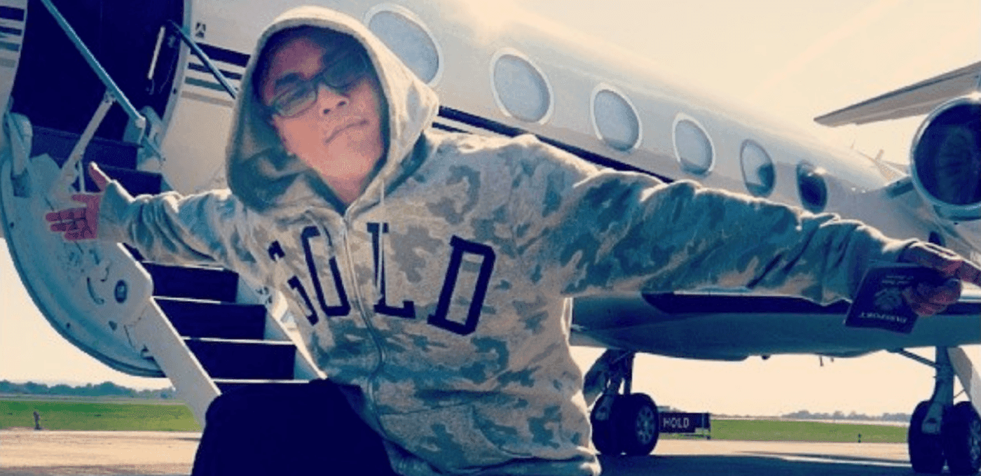 Person wearing a camo hoodie and glasses stands with arms outstretched beside a small private jet on the tarmac.