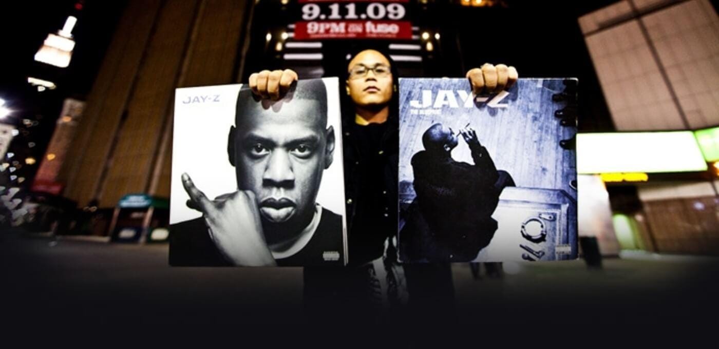 Person in a night city street holds two Jay-Z posters: left a black-and-white close-up, right a blue-toned pose image; blurred lights in the background.