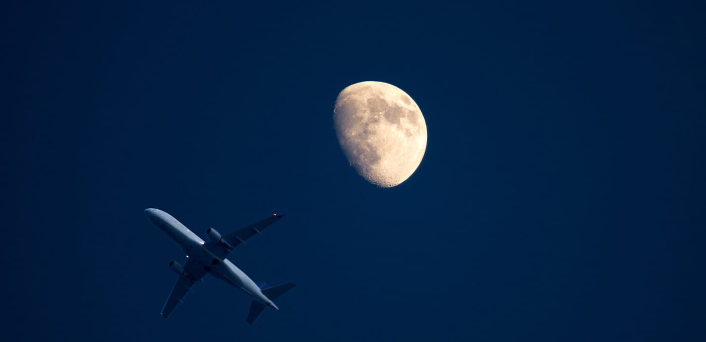Silhouette of a commercial airplane flying across a dark blue sky with a large illuminated Moon in the upper-right.