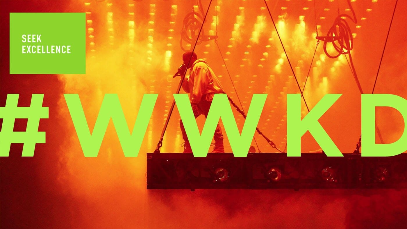 Performer on a suspended platform sings into a mic amid orange lights; lime text #WWKD spans the scene with a green SEEK EXCELLENCE box.