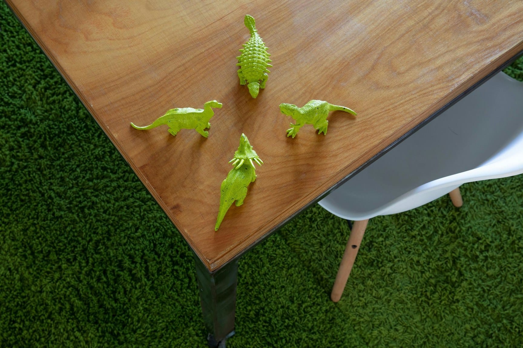 Four lime-green dinosaur figurines on a wooden table outdoors, with a white chair and green grass beneath.
