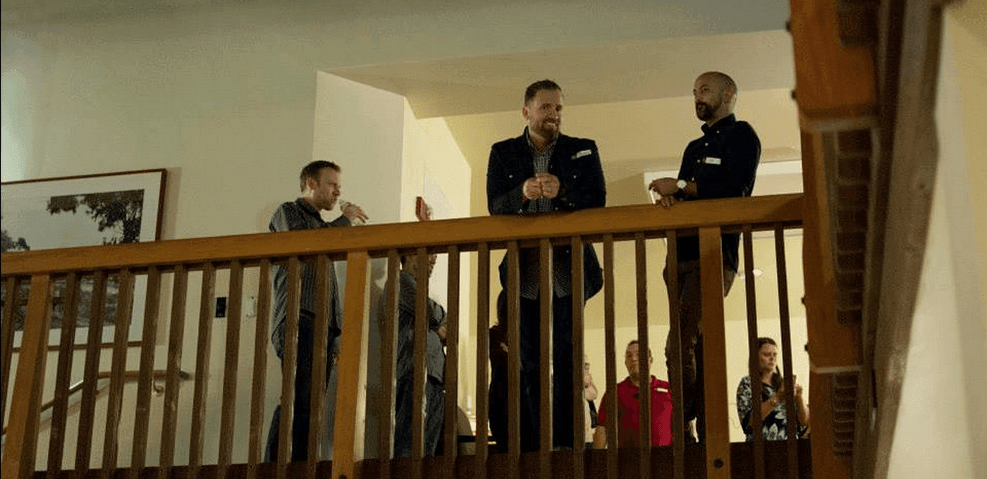 Group of people on an upstairs balcony, leaning on a wooden railing and conversing, while others below clap in a warmly lit home interior.