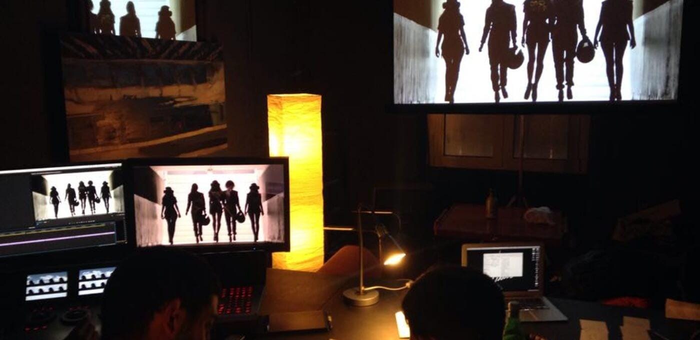 Dim control room with multiple screens displaying silhouettes of people walking; a tall yellow lamp illuminates a desk with editing gear.