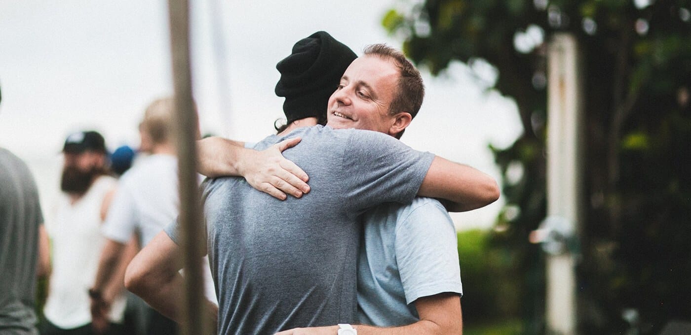 Two people share a warm hug outdoors, smiling; one wears a gray T-shirt and a black beanie, with bystanders blurred in the background.