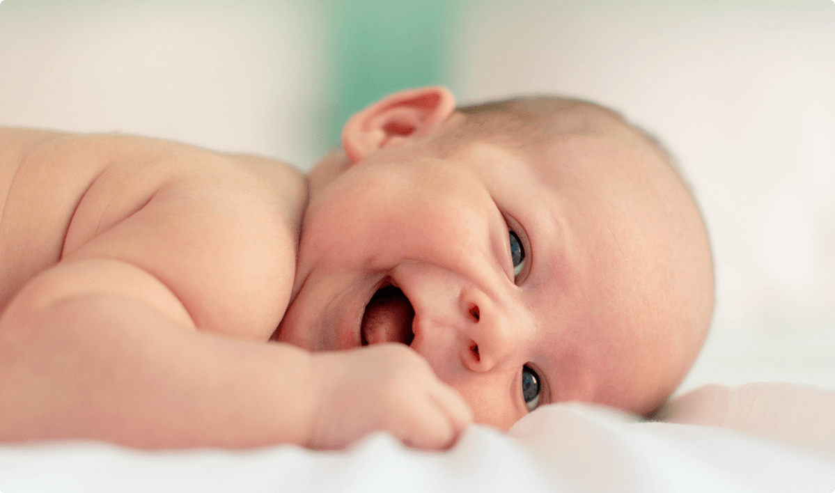 Newborn lying on a soft white surface, head turned toward the camera, mouth open in a smile, blue eyes and pink cheeks, pastel blurred background.