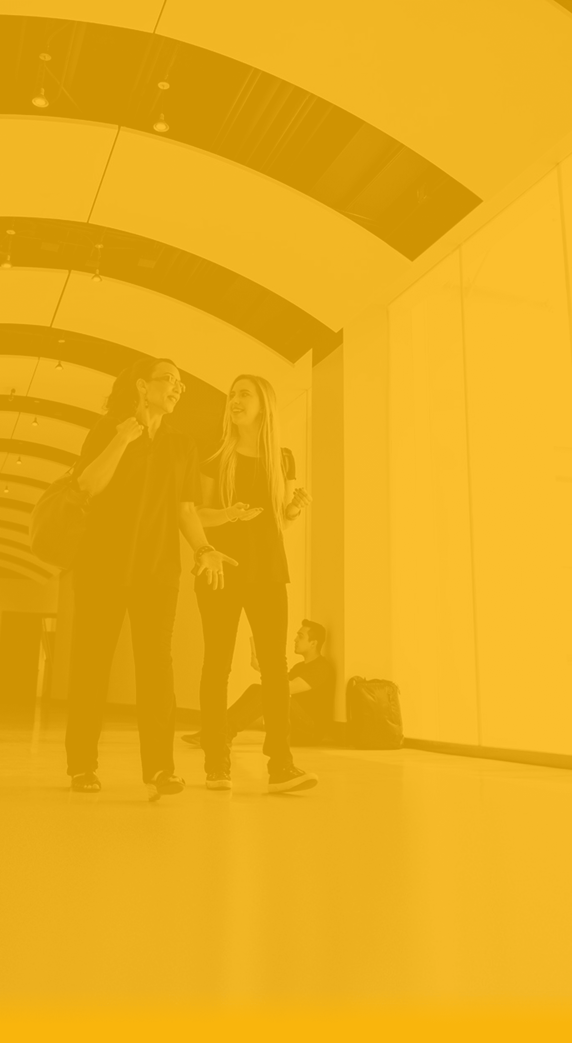 Two people stand in a bright hallway with curved ceiling panels, chatting and gesturing; a third person sits on the floor with a backpack in the background.
