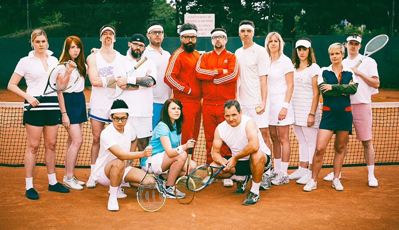 Group of people on a clay tennis court posing for a retro-style photo; many wear white outfits, two in red tracksuits center holding rackets.