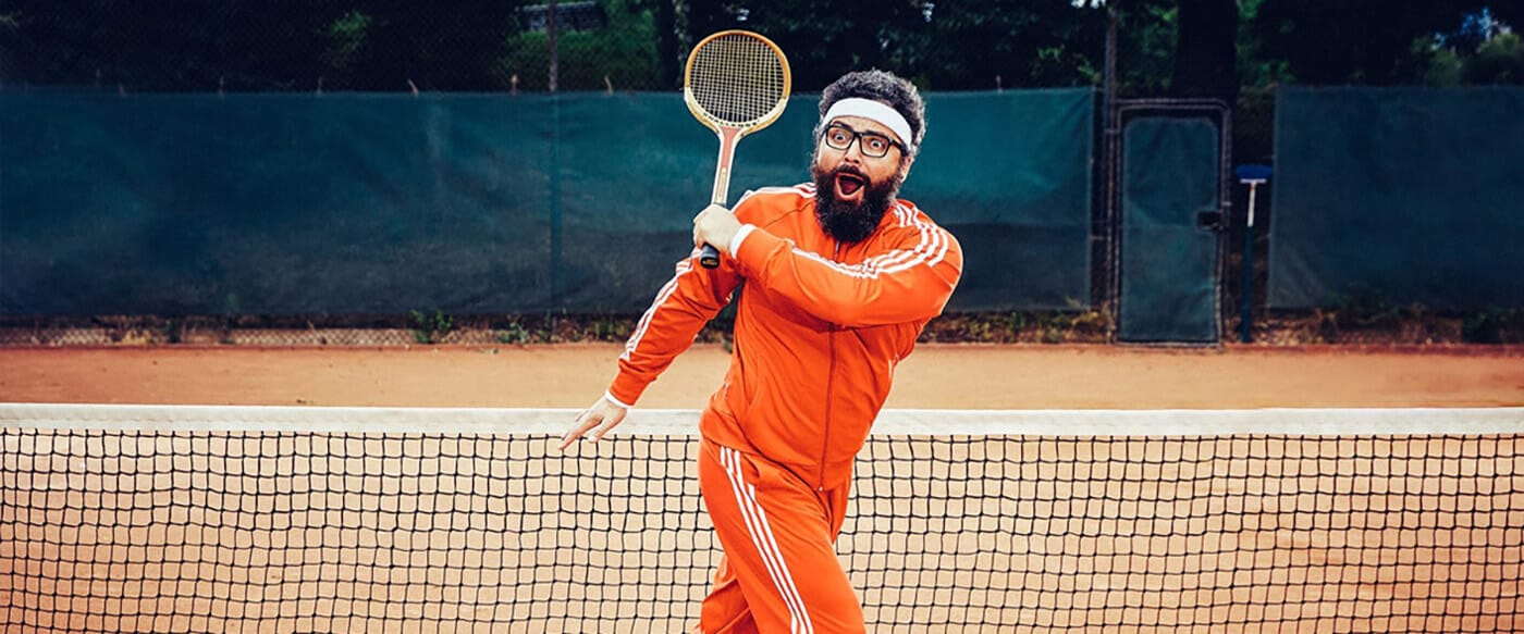Bearded person in an orange tracksuit with a white headband and glasses swings a tennis racket mid-swing on a clay court near the net.