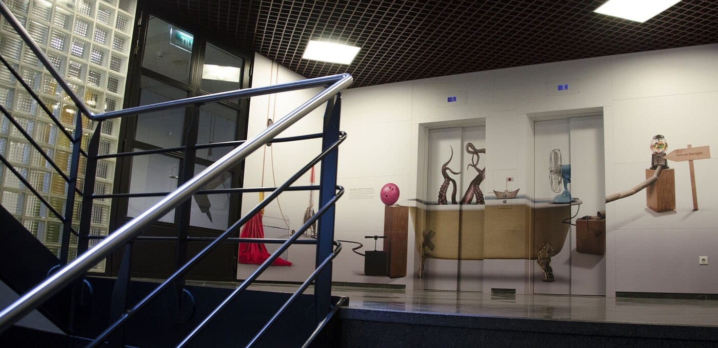 Indoor gallery with a metal railing in the foreground; mural shows a bathtub with legs and whimsical objects, pink ball on a pedestal, lights.