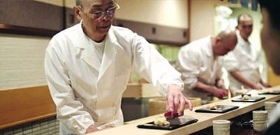 A person in a white chef coat and glasses plates food at a counter, with other chefs in white coats visible in the background.