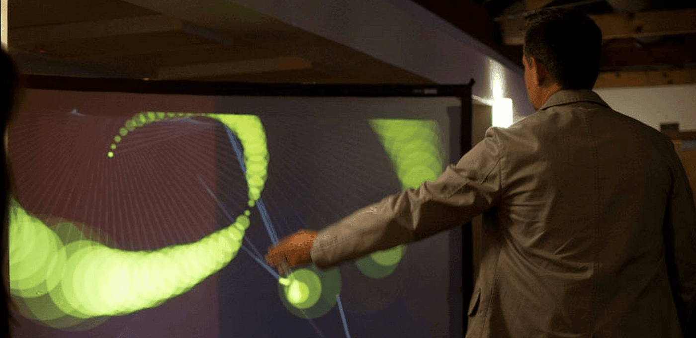 Person in a light blazer stands before a large interactive screen, touching glowing green circular patterns and blue lines projected across display.