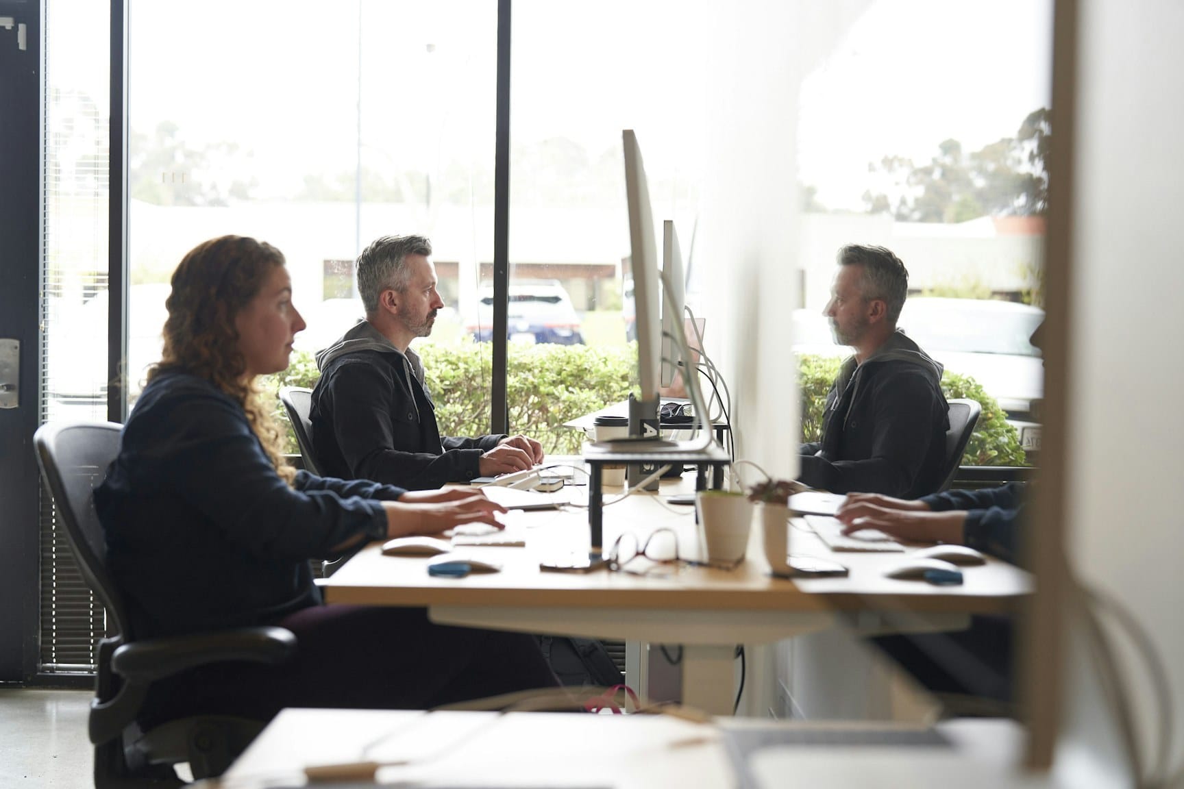 Three people sit at a long, bright office desk with computer monitors; large windows behind them, plants on the desk, and an outdoor view.