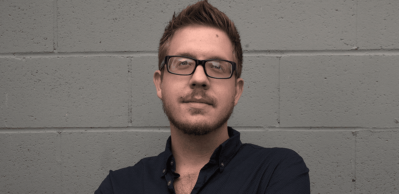 Person with short brown hair and glasses, wearing a dark button-down shirt, stands against a gray concrete block wall.