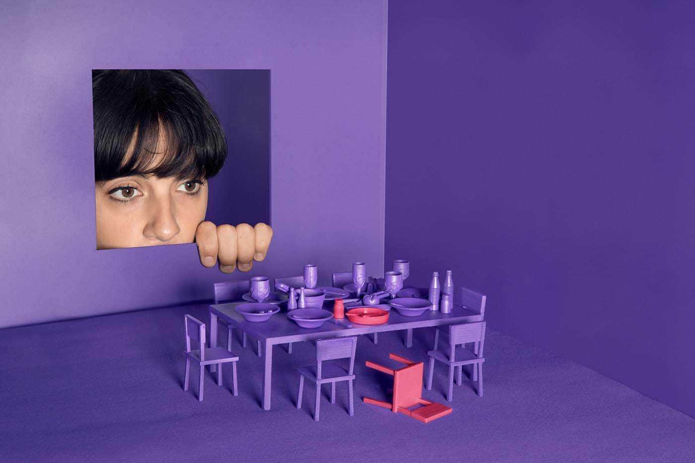 Person peeks through a square cutout in a purple wall; a purple dining table with matching chairs sits in the room, with an orange chair on the floor.