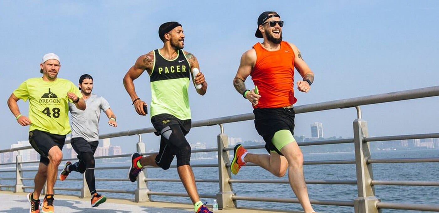 Four runners on a seaside boardwalk, wearing bright athletic gear, smiling as they sprint past a railing with a city skyline in the distance.