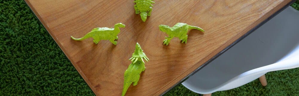 From above, a wooden table with four bright green toy dinosaurs arranged around its corner, on a green lawn with a white chair leg visible.