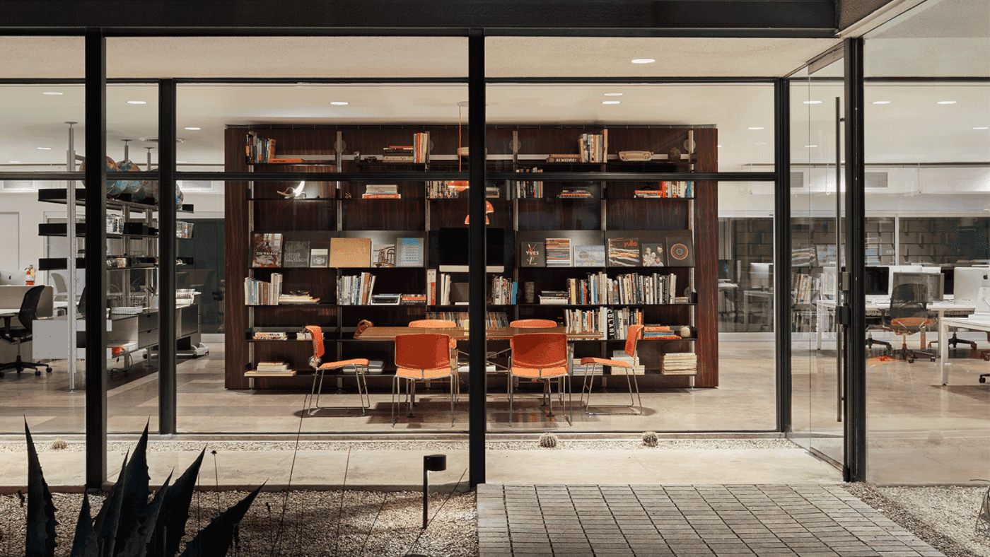 Glass-walled reading room with a dark wood bookshelf wall, orange chairs around a table, and a distant office area visible through the glass.