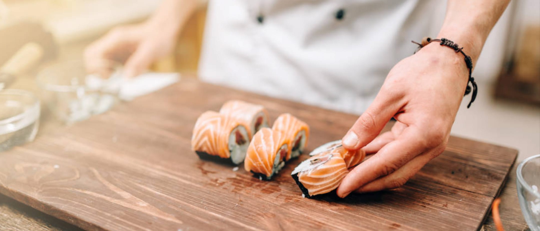 Hand with bracelet slicing salmon sushi rolls on a wooden cutting board; several pieces arranged, background blurred.
