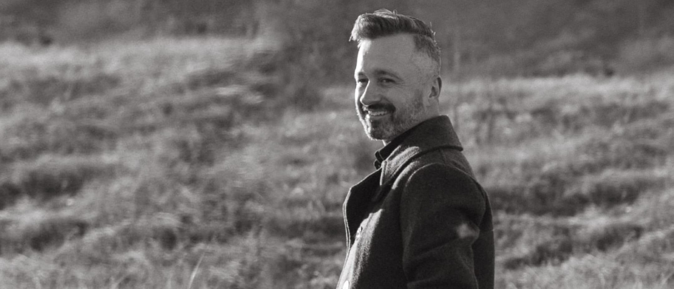 A person with short hair and a beard stands in a grassy field, wearing a dark coat, smiling and looking to the right.