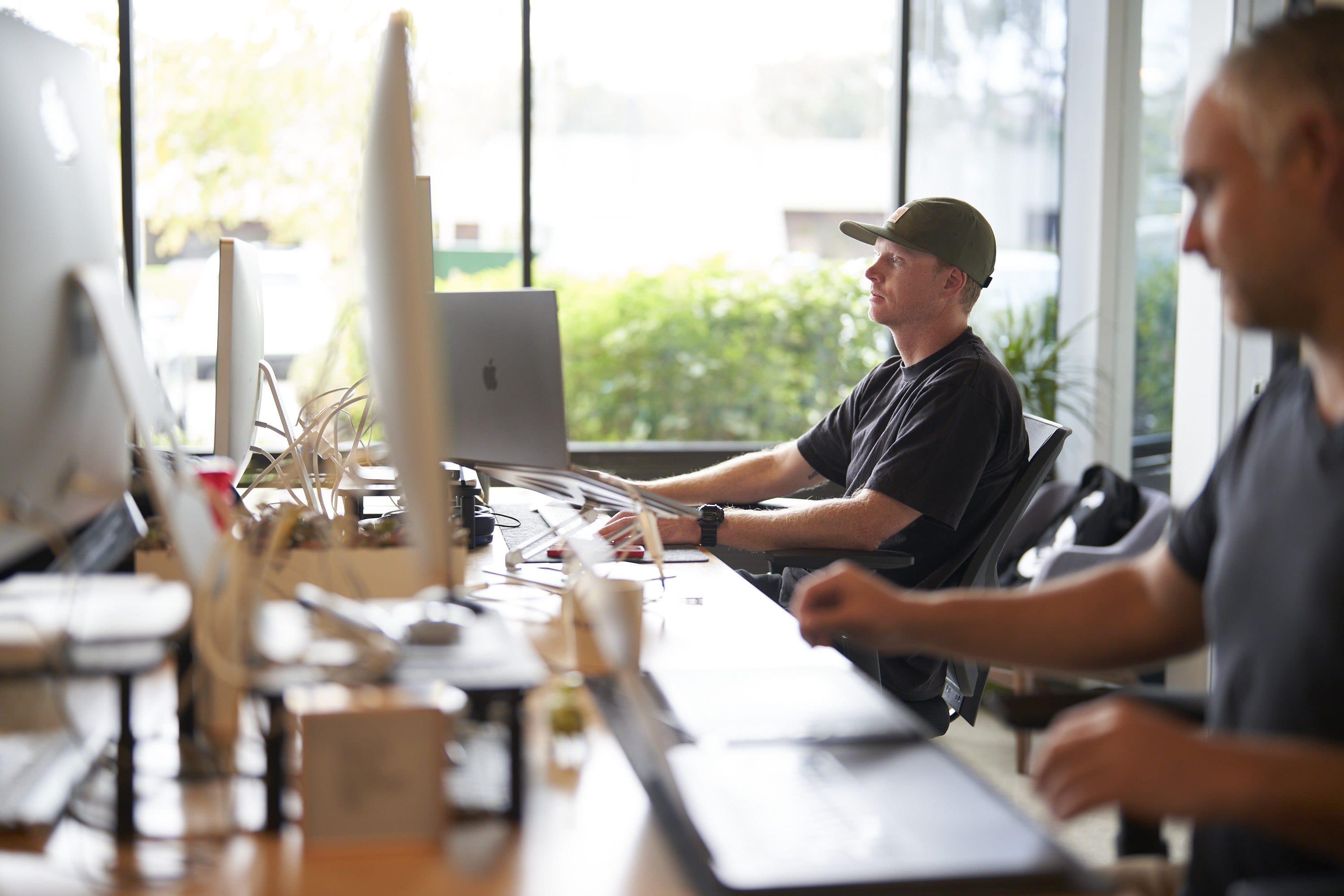 Open-plan office with two people at desks by large windows; foreground person is blurred, the other wears a cap and focuses on a computer among plants and cables.