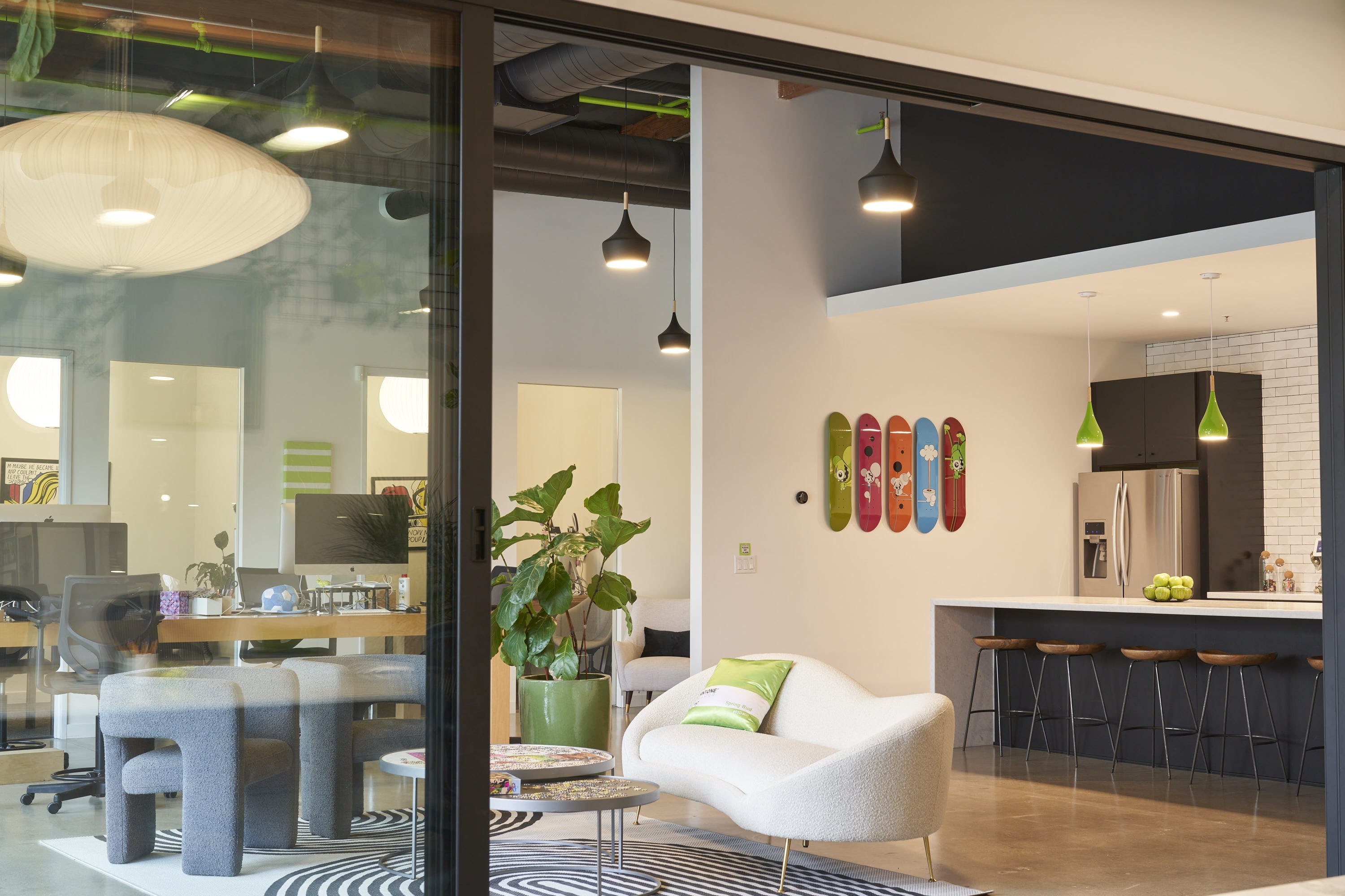 Open-plan workspace with glass walls, a white curved sofa with a green cushion, colorful skateboard wall art, and a kitchen island with stools.