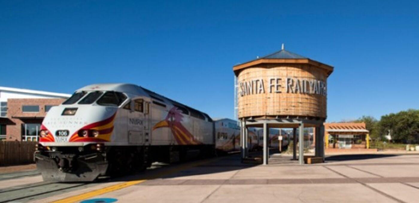 White passenger train with red and gold accents at a platform beside a wooden water tower labeled Santa Fe Railway under a clear blue sky.