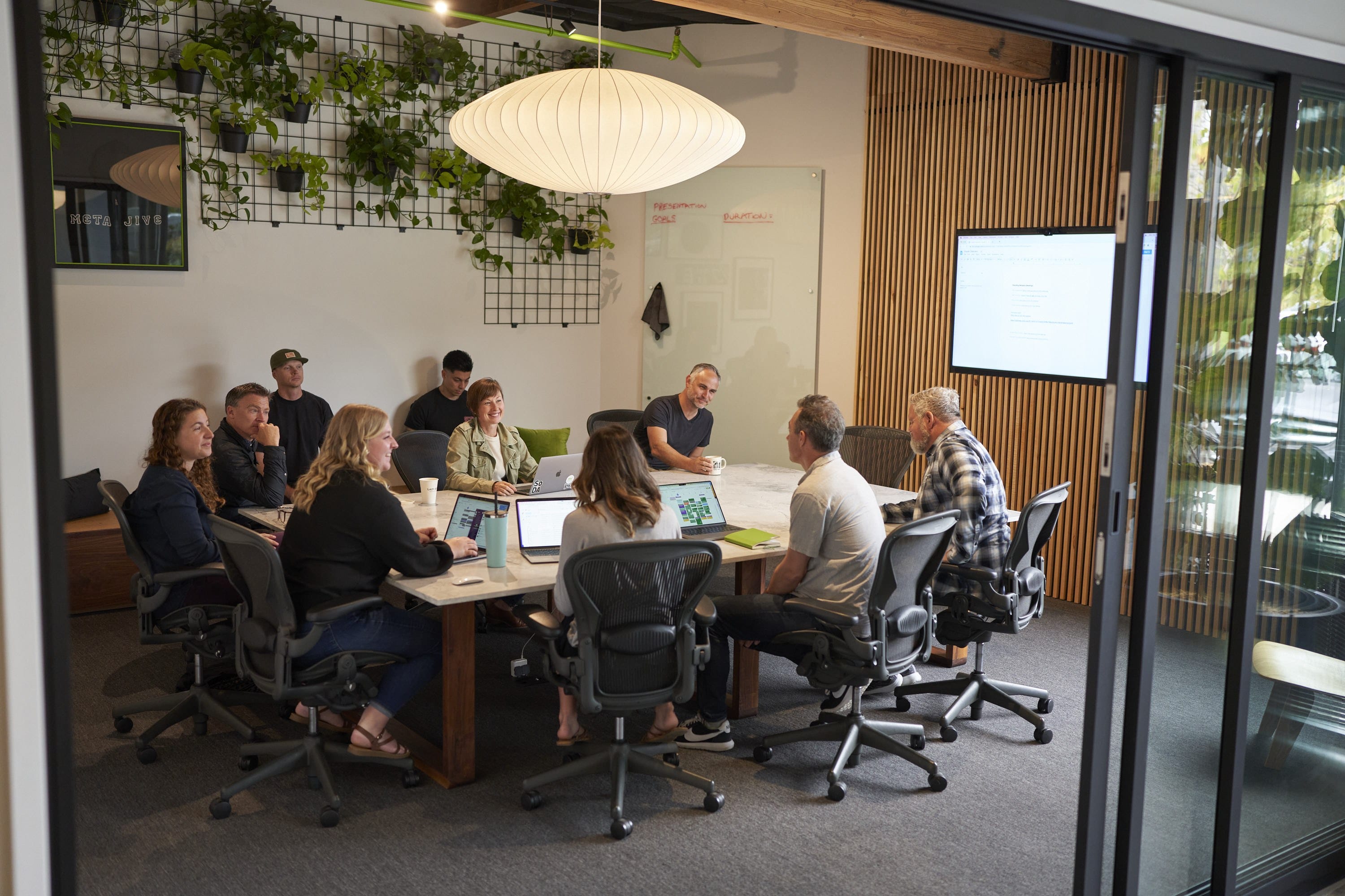 Group of diverse people seated around a conference table in a modern meeting room with laptops, a whiteboard, plants, and a hanging light.