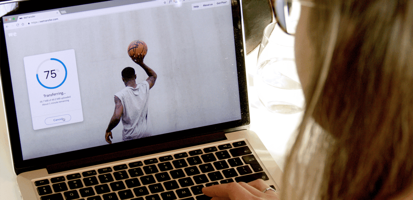 Laptop displays a 75% transfer progress dialog beside a basketball player lifting a ball overhead; a person with long hair types at the keyboard.