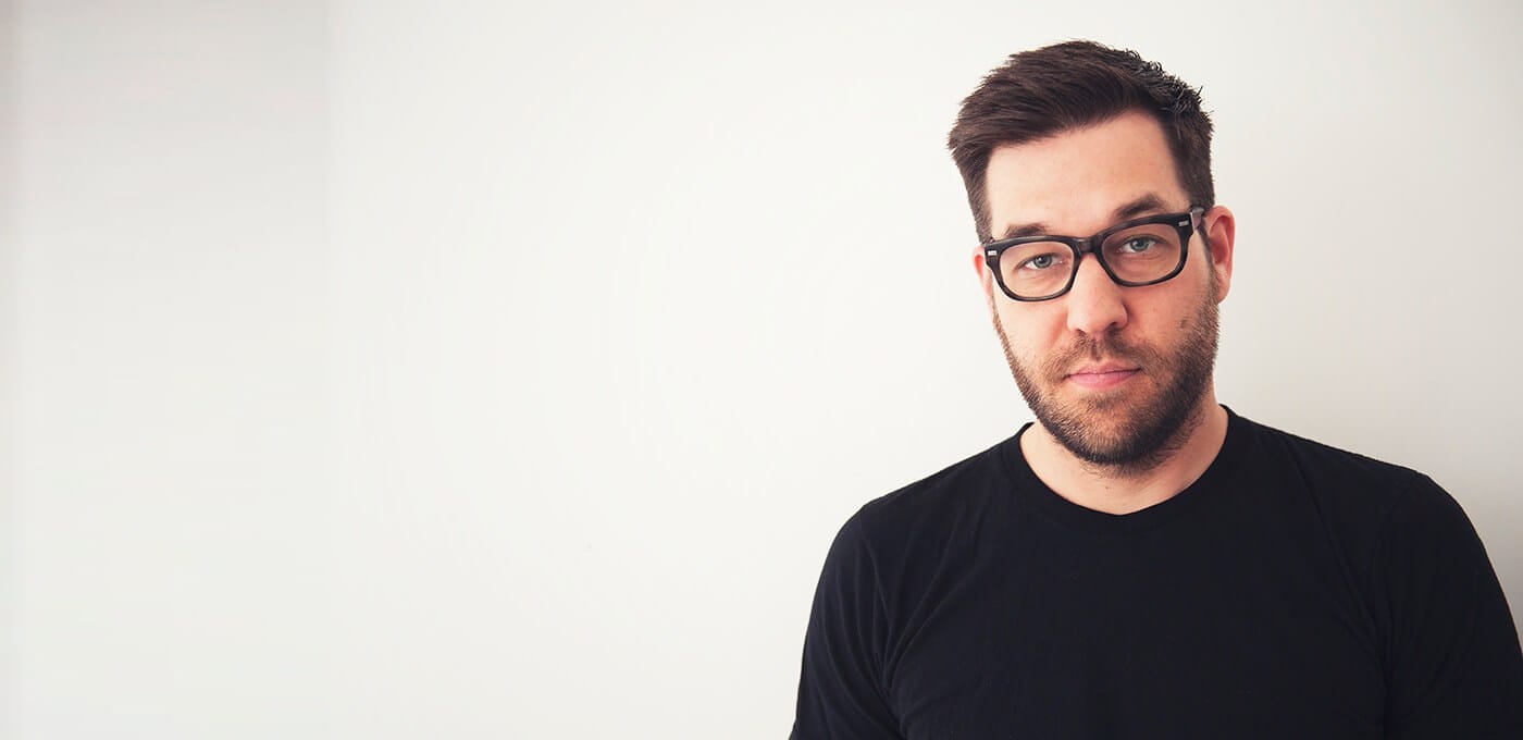A person with short dark hair and beard, wearing black-framed glasses and a black shirt, gazing at the camera against a light background.