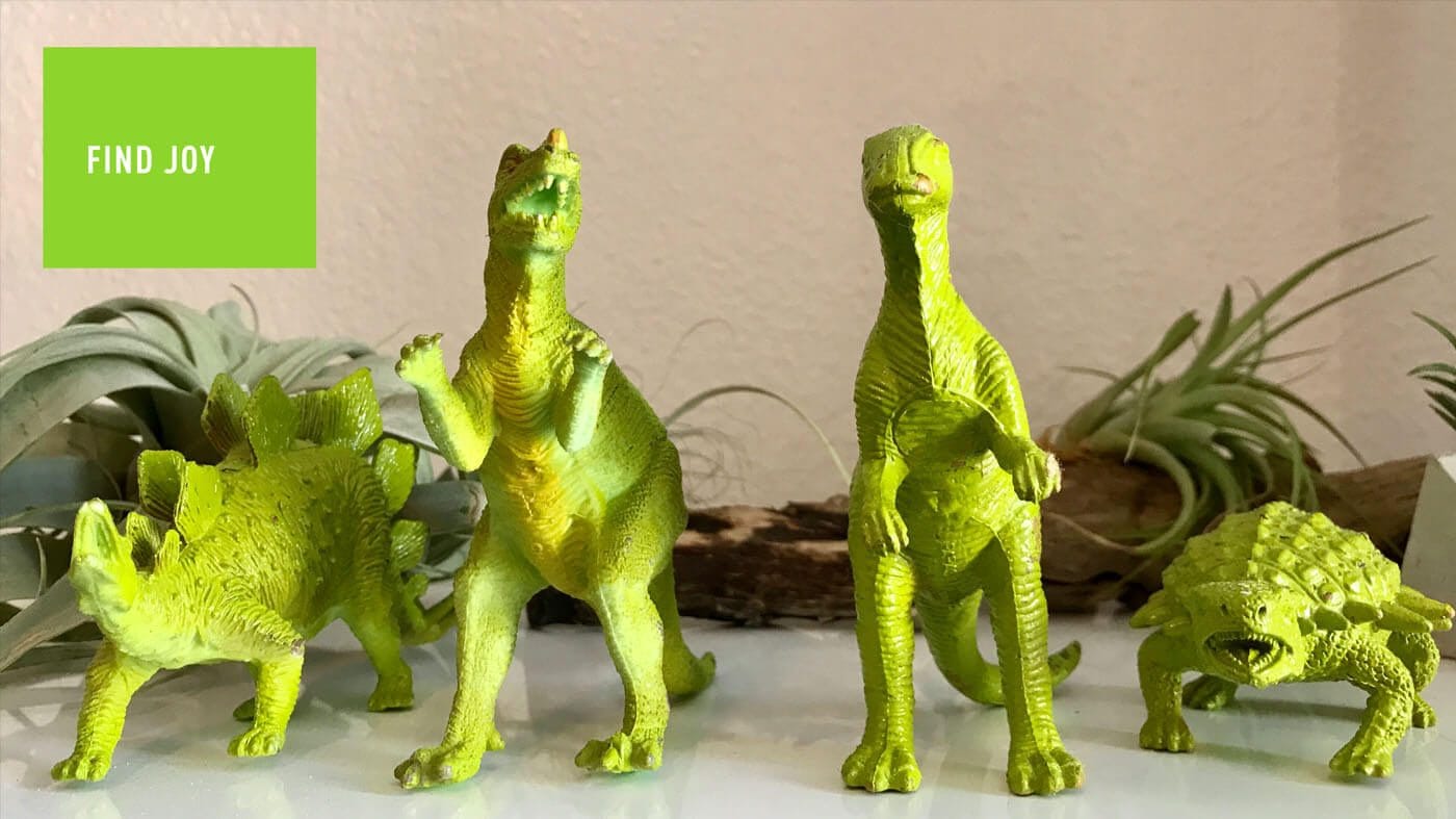 Neon lime-green dinosaur figurines stand on a white surface with plants in the background; a lime square reads 'FIND JOY' in the top left.